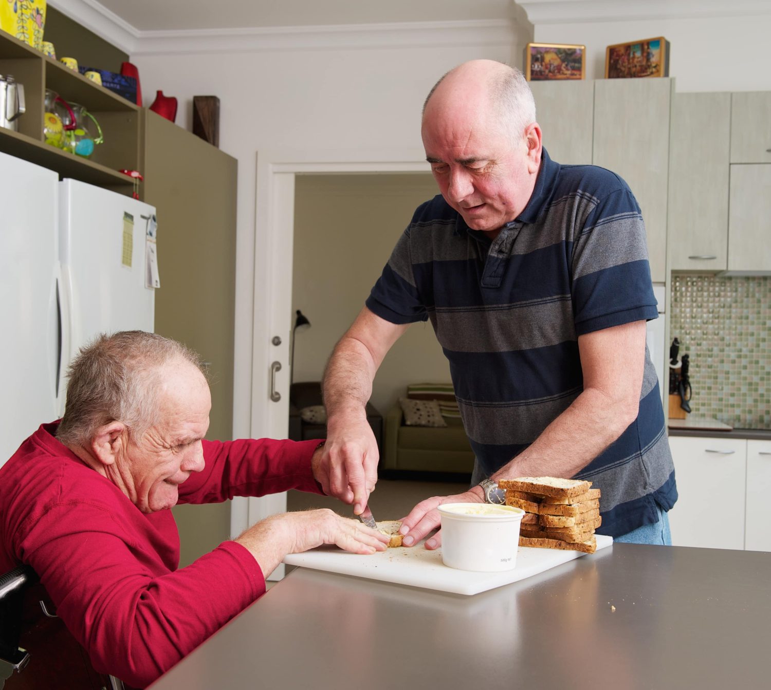 Male home Carer assisting senior Man with a Disability in a kitchen.